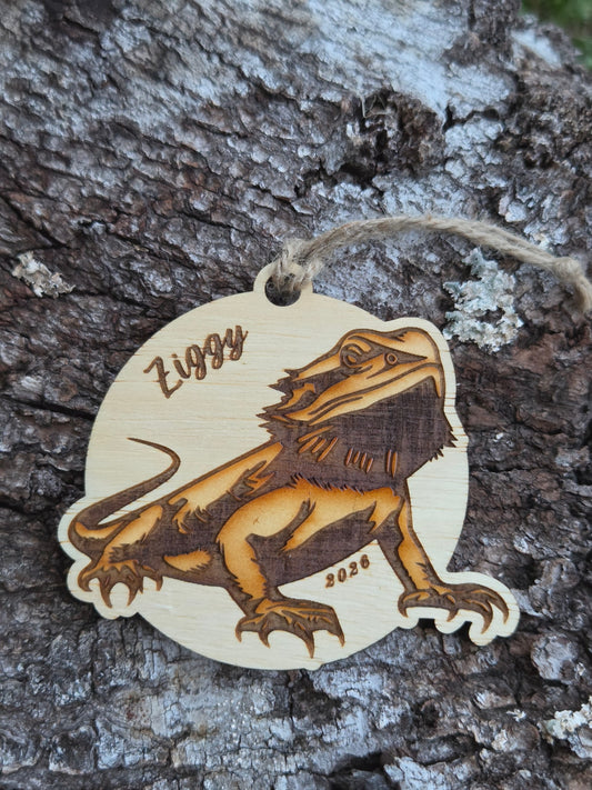 Wooden bearded dragon ornament on a round, with a sample name and year engraved, resting on tree bark