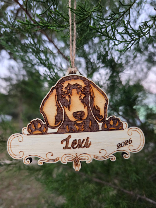 Wooden dachshund ornament, paws over a plaque engraved Lexi and 2026, hanging on an evergreen branch