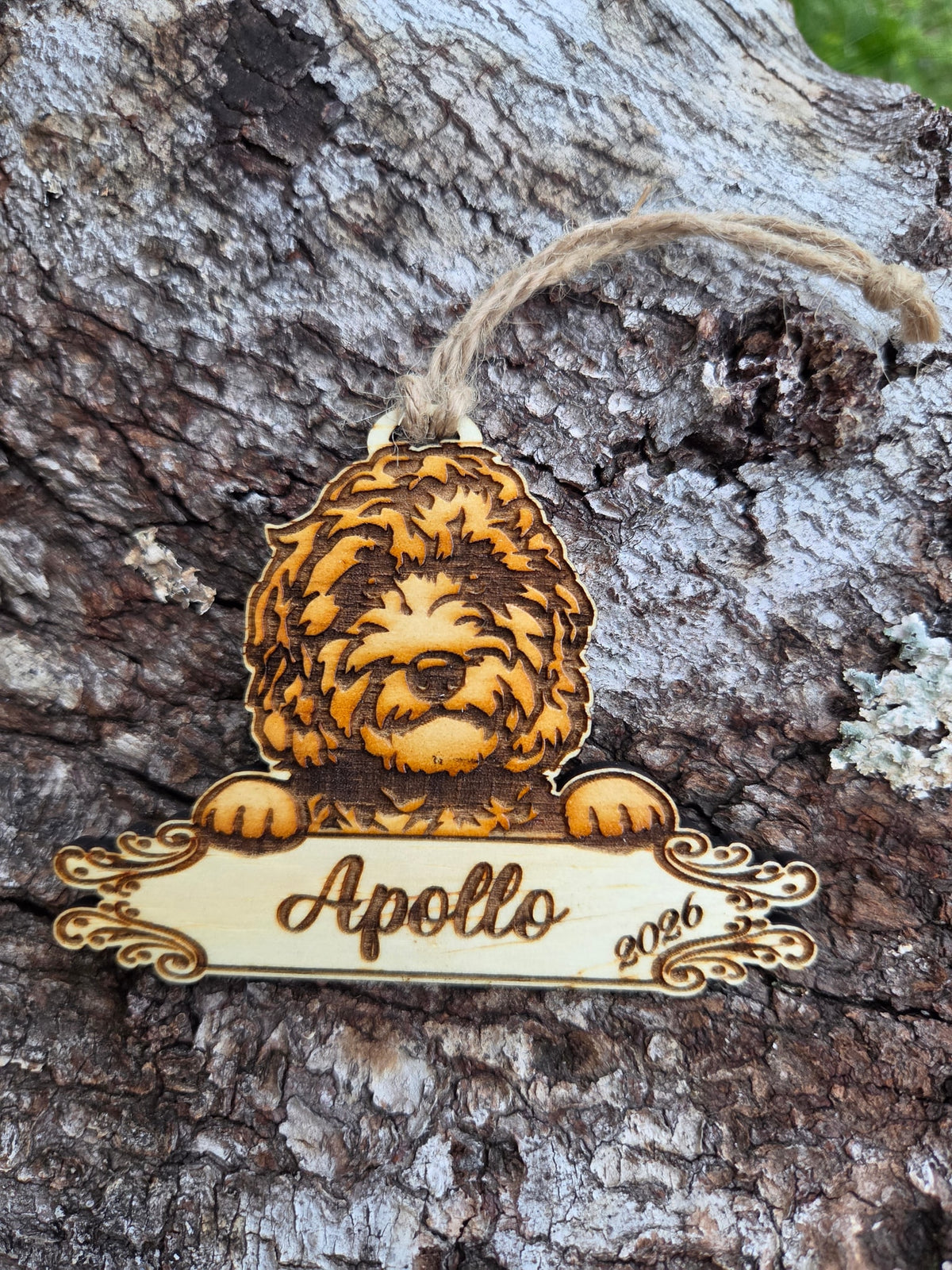 Wooden Doodle dog ornament, paws over a plaque engraved with a sample name and the year, resting on tree bark