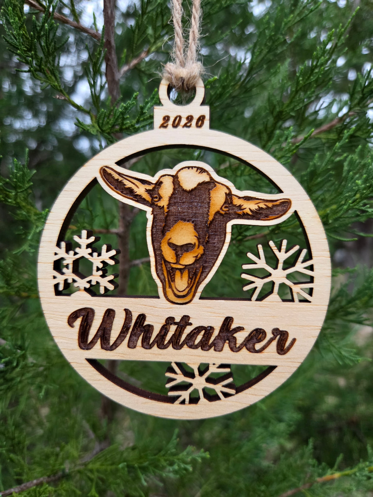 Goat face and snowflakes ornament hanging by its twine loop on an evergreen branch