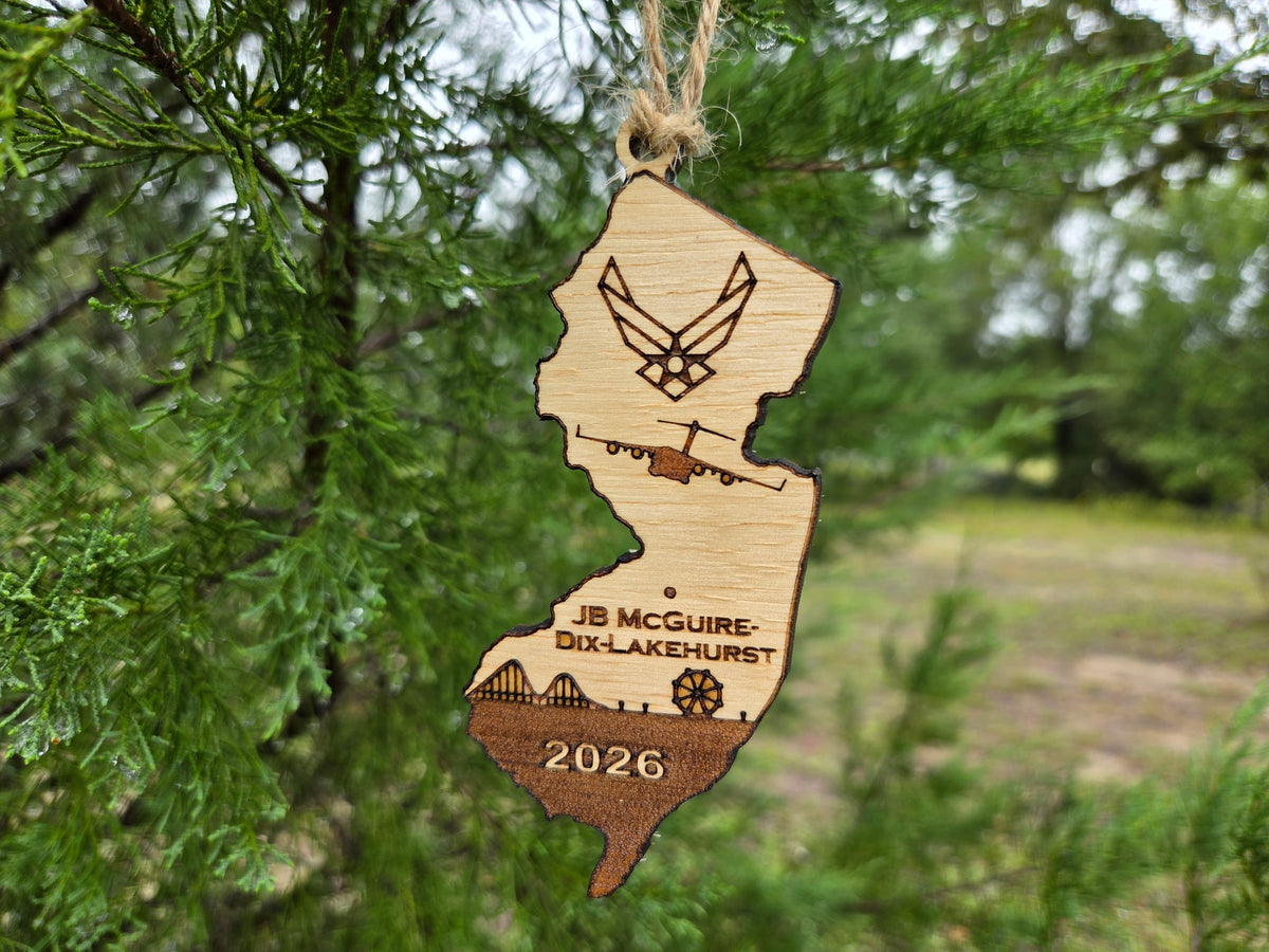 JB McGuire-Dix-Lakehurst ornament hanging on an evergreen, with a field behind