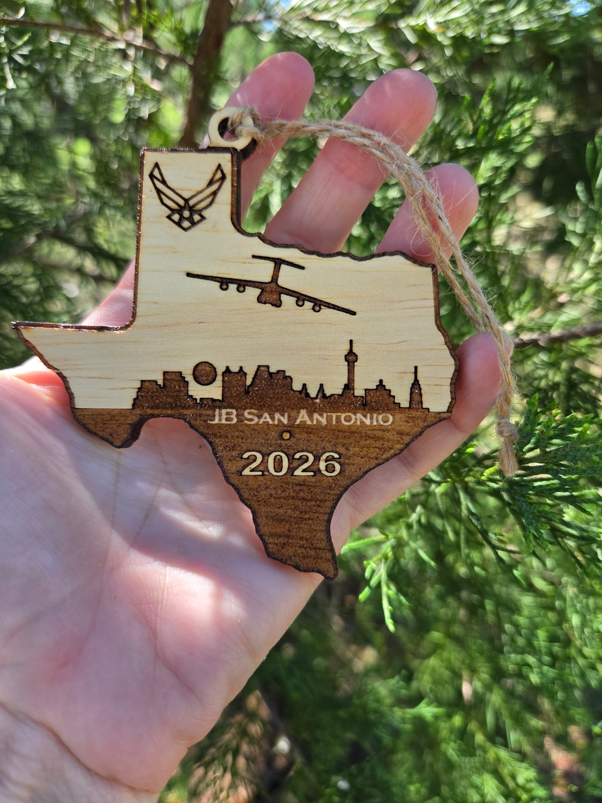 JB San Antonio Texas ornament held up in an open hand in front of an evergreen