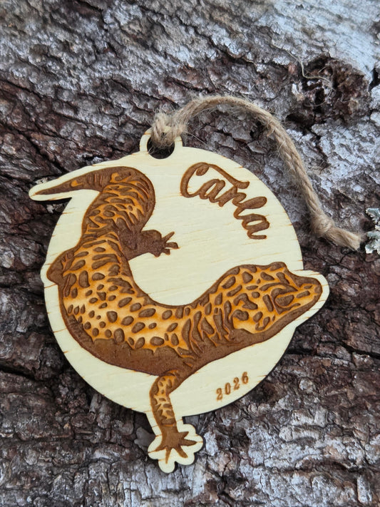 Wooden leopard gecko ornament with a sample name and year engraved, resting on tree bark