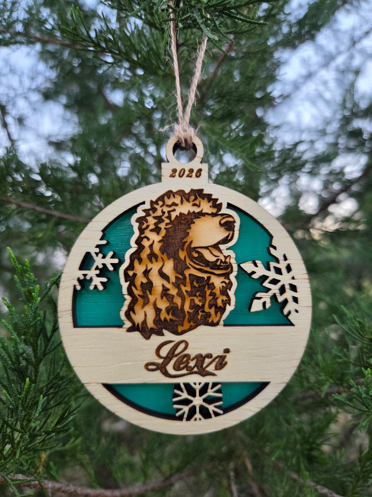Round wooden poodle head ornament with a teal back, engraved Lexi and 2026, hanging on an evergreen branch