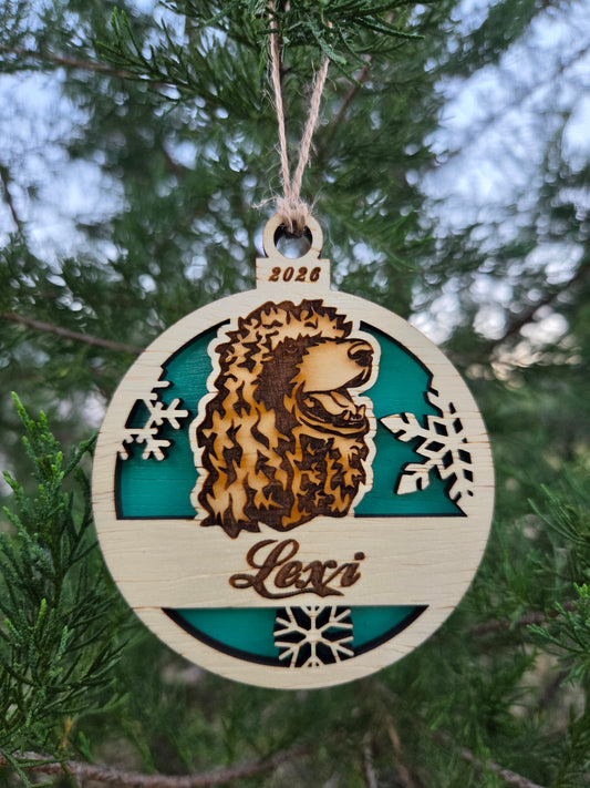 Round wooden poodle head ornament with a teal back, engraved Lexi and 2026, hanging on an evergreen branch