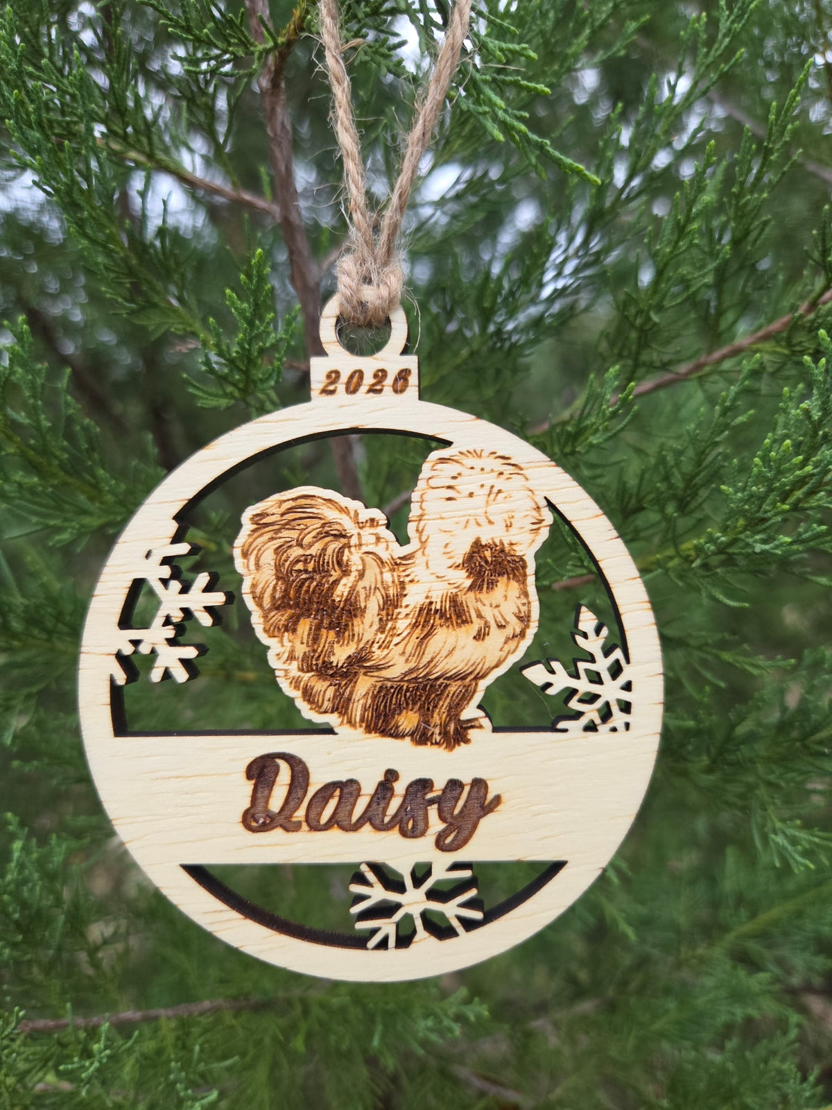 Silkie chicken and snowflakes ornament hanging by its twine loop on an evergreen branch