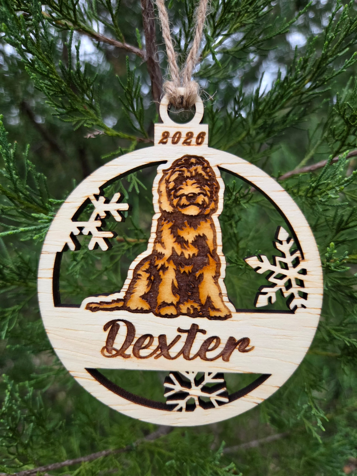 Sitting doodle dog and snowflakes ornament hanging by its twine loop on an evergreen branch