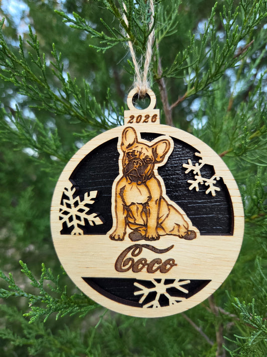 Sitting French Bulldog and snowflakes ornament with a black back, engraved Coco and 2026, hanging on an evergreen