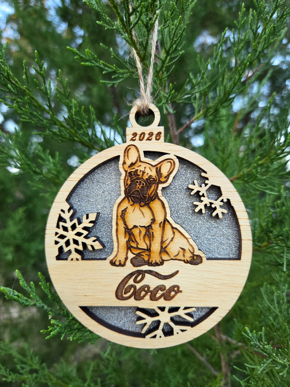 Sitting French Bulldog and snowflakes ornament with a silver glitter back, engraved Coco and 2026, on an evergreen