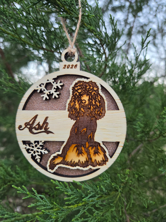 Round wooden sitting poodle ornament with a rose gold glitter back, engraved Ash and 2026, on an evergreen