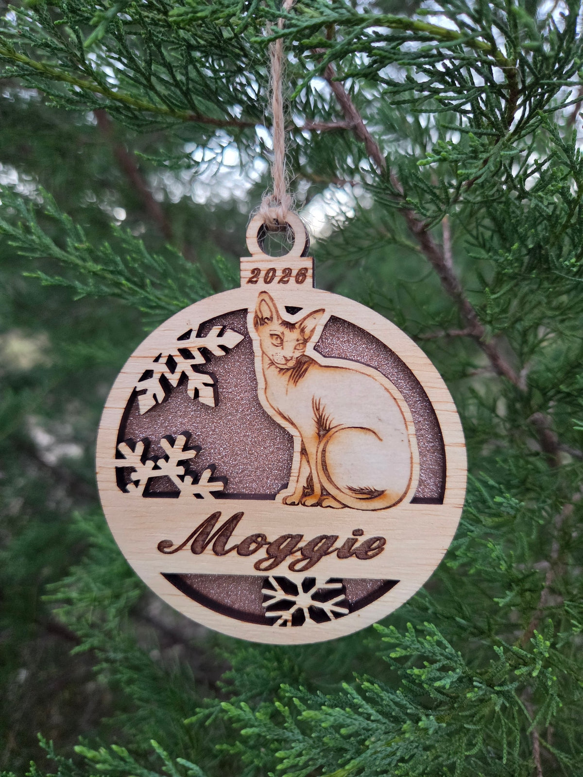 Sitting Sphynx cat ornament with a rose gold glitter back, engraved Moggie and 2026, hanging on an evergreen