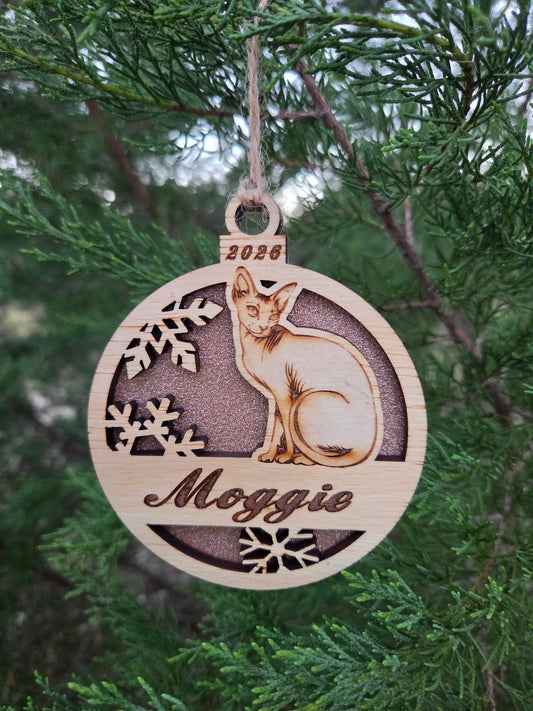 Sitting Sphynx cat ornament with a rose gold glitter back, engraved Moggie and 2026, hanging on an evergreen