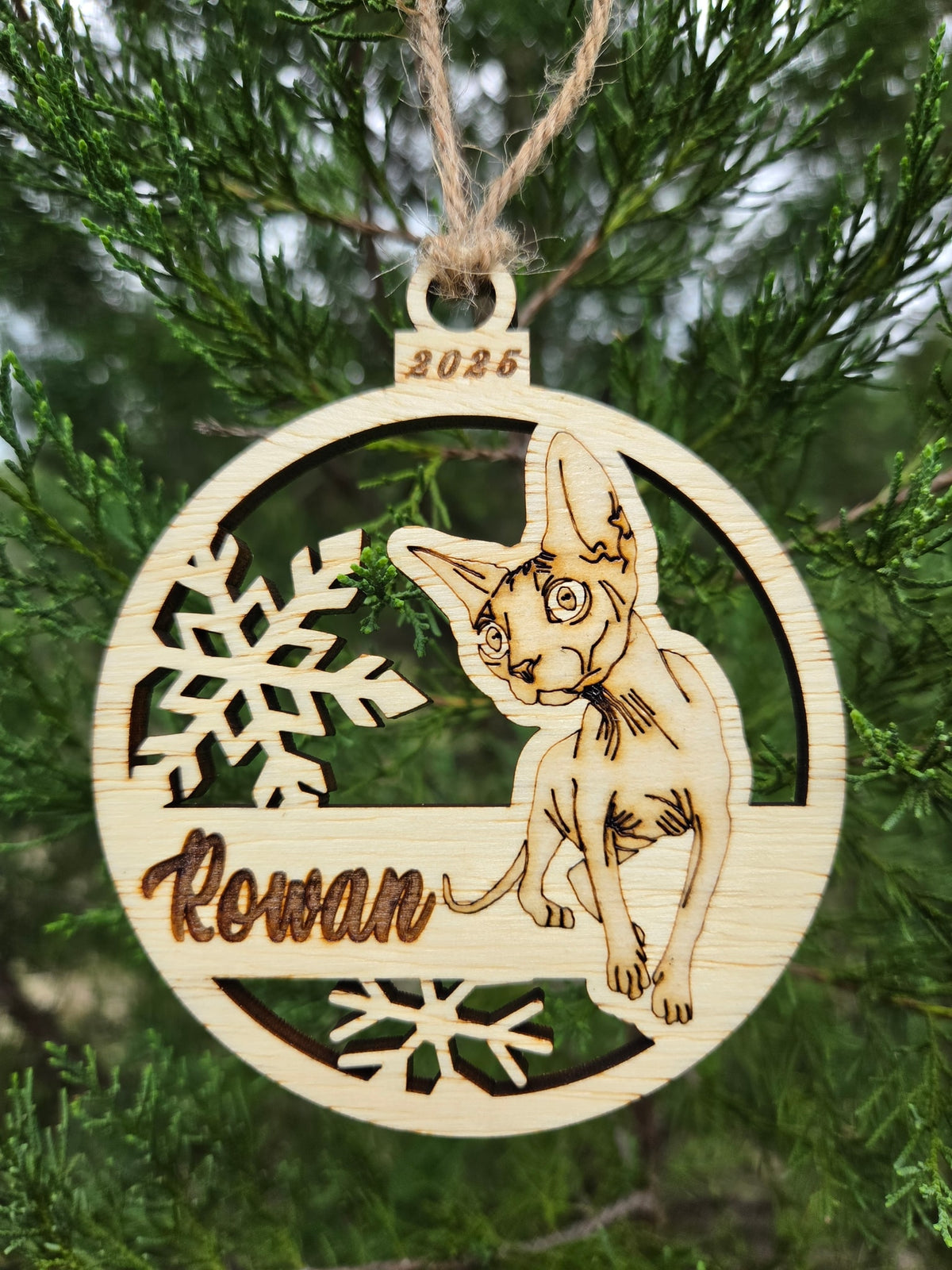 Sphynx cat and snowflakes ornament hanging by its twine loop on an evergreen branch