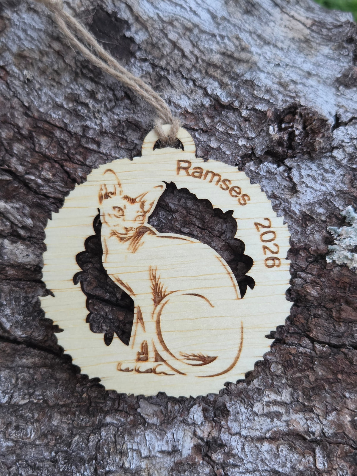 Wooden Sphynx cat in a wreath ornament, a sample name and year along the top, resting on tree bark