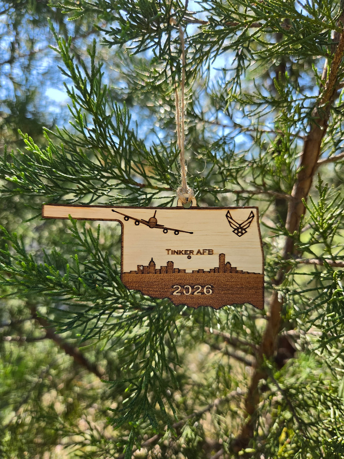 Oklahoma-shaped wooden ornament engraved with Tinker AFB, an E-6B, a skyline and the year, hanging on an evergreen