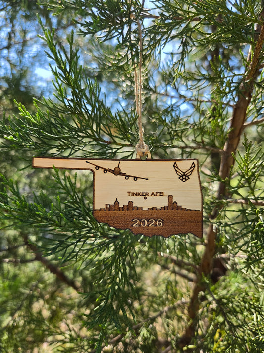Oklahoma-shaped wooden ornament engraved with Tinker AFB, an E-6B, a skyline and the year, hanging on an evergreen