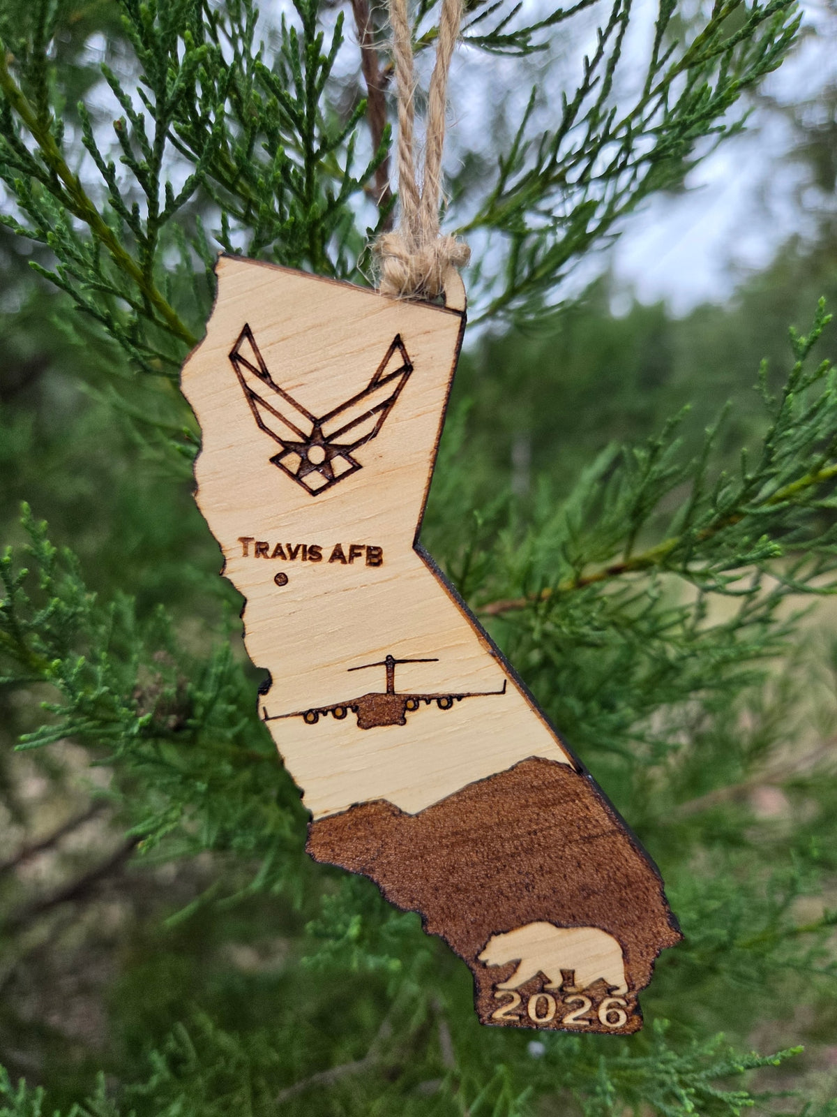 California-shaped wooden ornament engraved with Travis AFB, a C-17 and the year, hanging on an evergreen branch