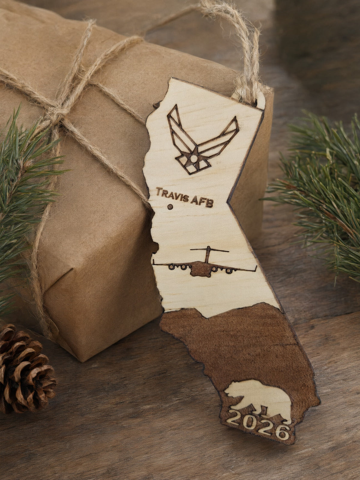 Travis AFB California ornament resting on a kraft paper gift tied with twine, beside pine sprigs and a pine cone